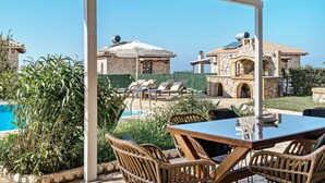 A covered outdoor seating area with a pool, wicker furniture, and stone buildings in the background. - "Villa Alexandros" with Sea View, Private Pool & Wi-Fi (Lefkada)