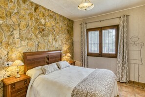 Ein Schlafzimmer mit Steinwand, einem hölzernen Kopfteil, einem Bett mit gemusterten Betttextilien, zwei Nachttischlampen, einem Fenster mit Vorhängen und einer Wandleuchte.