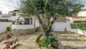 A stone-paved courtyard with a large tree, a white building with a blue door, and a stone wall.
