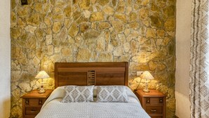A bedroom with a stone wall, a wooden headboard, a bed with patterned bedding, two bedside tables with lamps, and a curtain on the window.