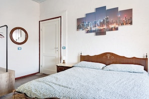 A bedroom with a wooden headboard, a bed with a patterned bedspread, a bedside table with a lamp, a wall-mounted mirror, and a cityscape artwork.