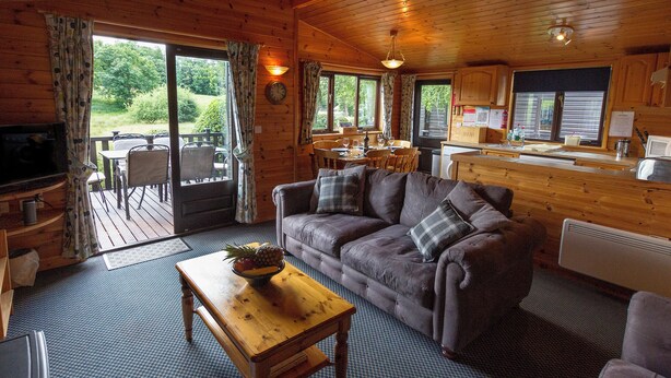 A cozy cabin interior with a wooden ceiling, a sofa, a coffee table with fruit, a dining area, and a kitchen with a window. - Whiteside Lodge at Burnside Park (Keswick)