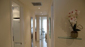 Un couloir avec un réfrigérateur blanc, une étagère en verre sur laquelle est posée une orchidée en pot, et une chaise au loin.