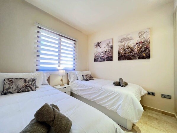 A hotel room with two beds, a window with blinds, a nightstand with a lamp, and two wall-mounted paintings.