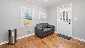 A room with a grey sofa, a white door, and a window with blinds. - Private 1BR Cottage In Downtown Historic Norcross (Norcross)