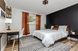 Ein Schlafzimmer mit einem Bett, einem Schreibtisch, einem Stuhl, einem Fenster mit Vorhängen und einem Nachttisch mit Pflanze.
