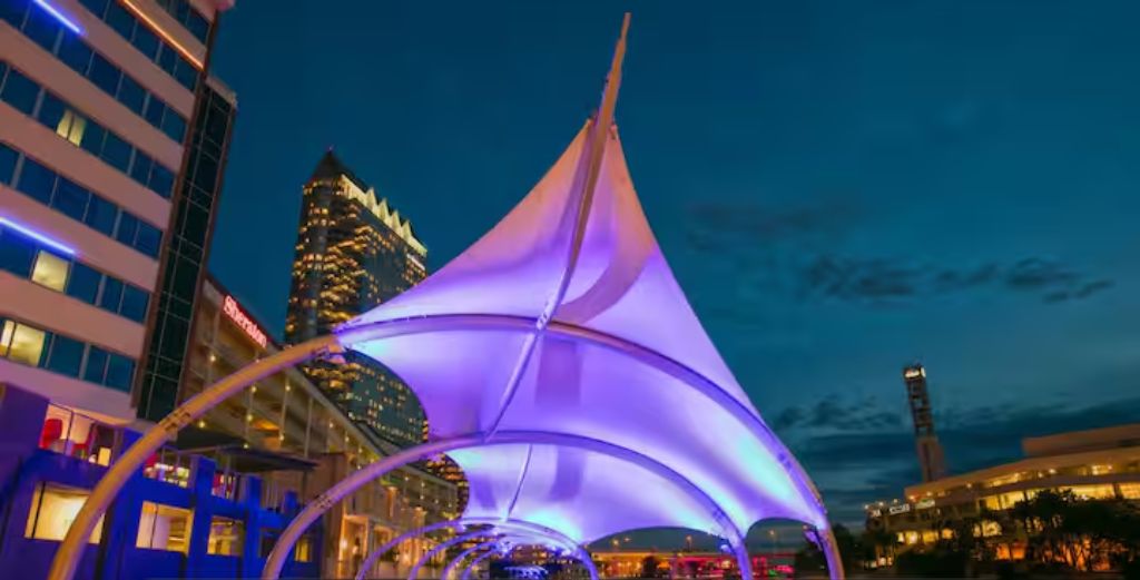 A nighttime view of the Tampa Riverwalk with a sail-like structure in the foreground and buildings in the background.