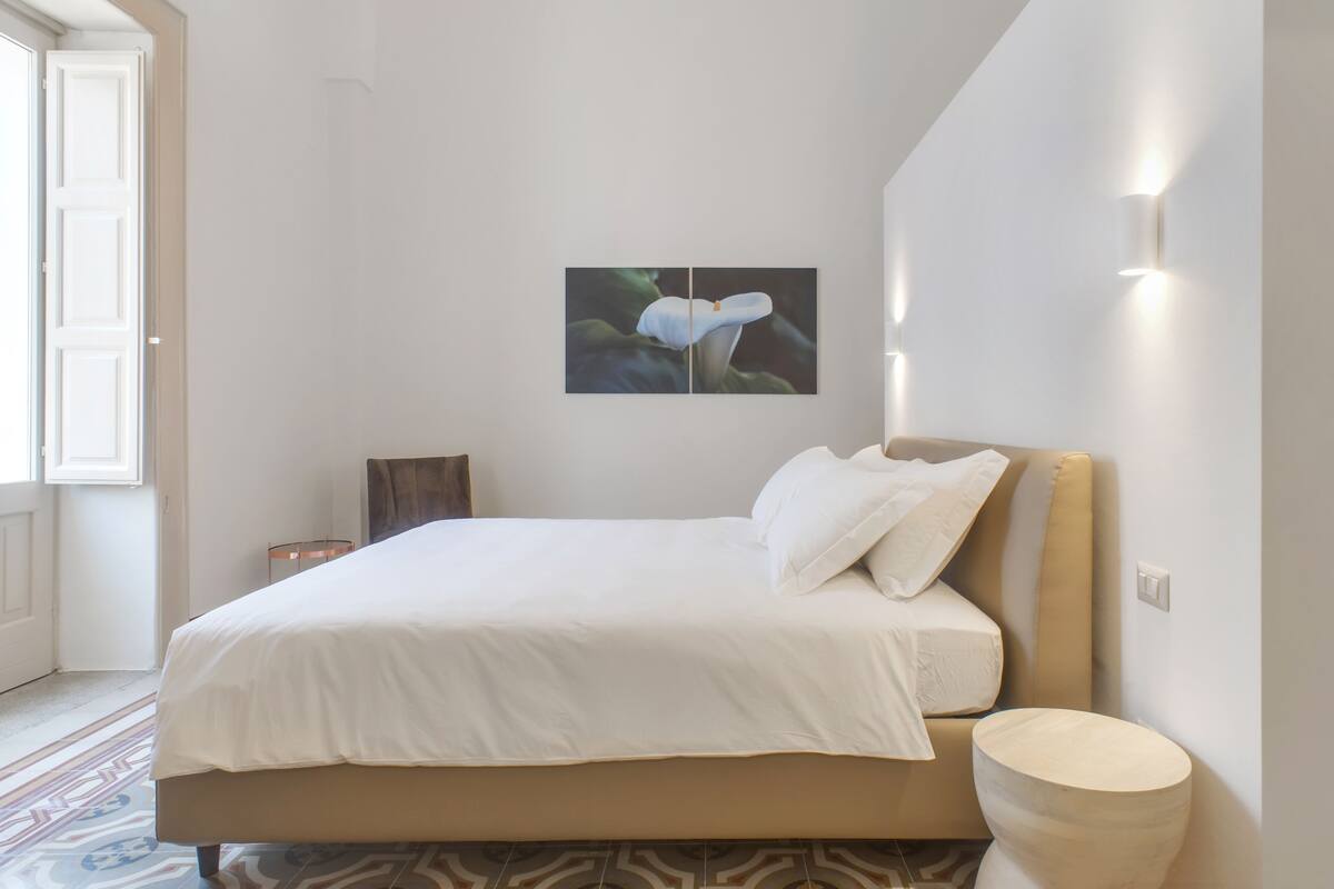 A neatly made bed with white linens, a wooden headboard, and a wall-mounted light fixture.