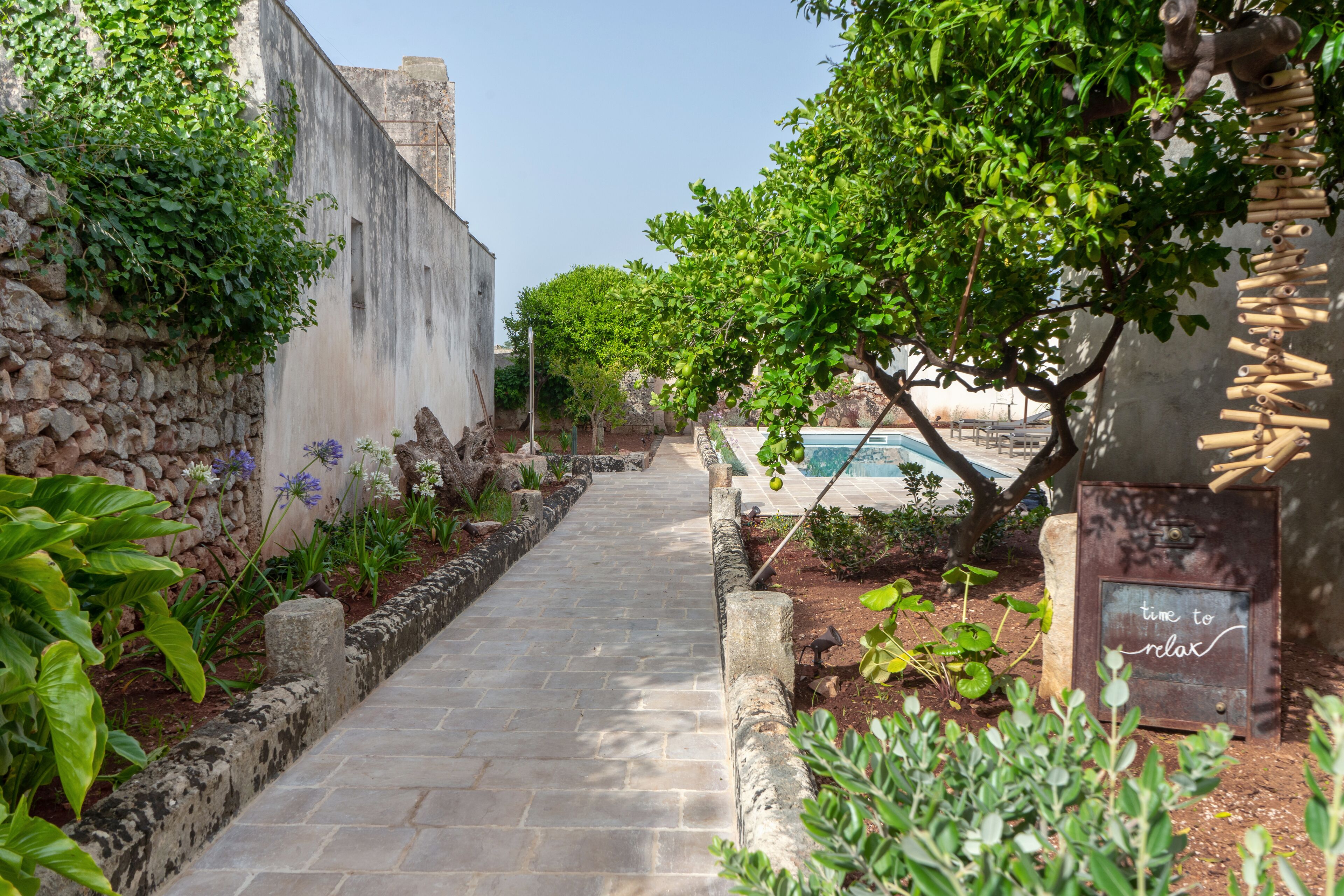 A stone pathway leading to a pool area with a rustic wall and greenery on one side.
