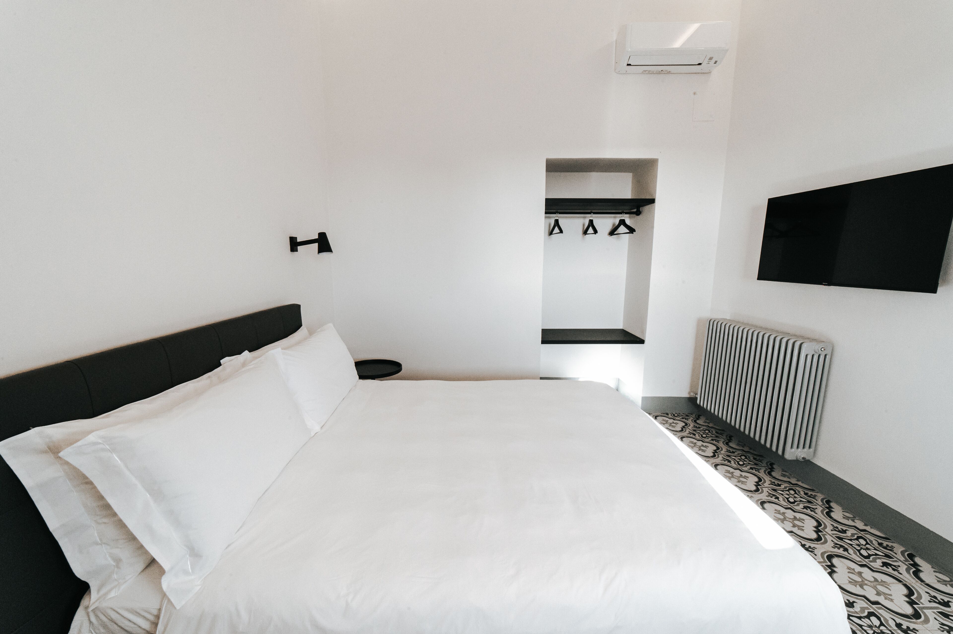 A hotel room with a large bed, a flat-screen TV mounted on the wall, a radiator, and a small shelf with hangers.