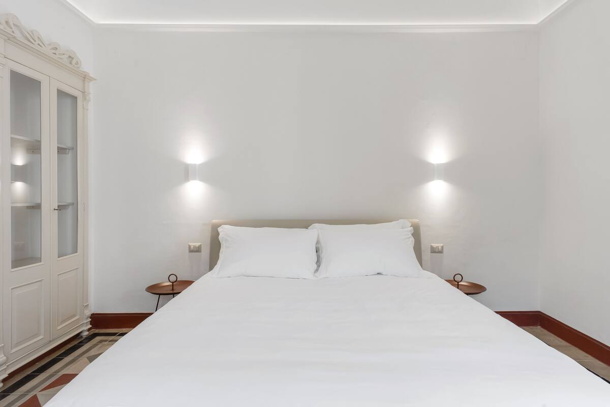 A neatly made bed with white linens in a room with white walls and a tiled floor.
