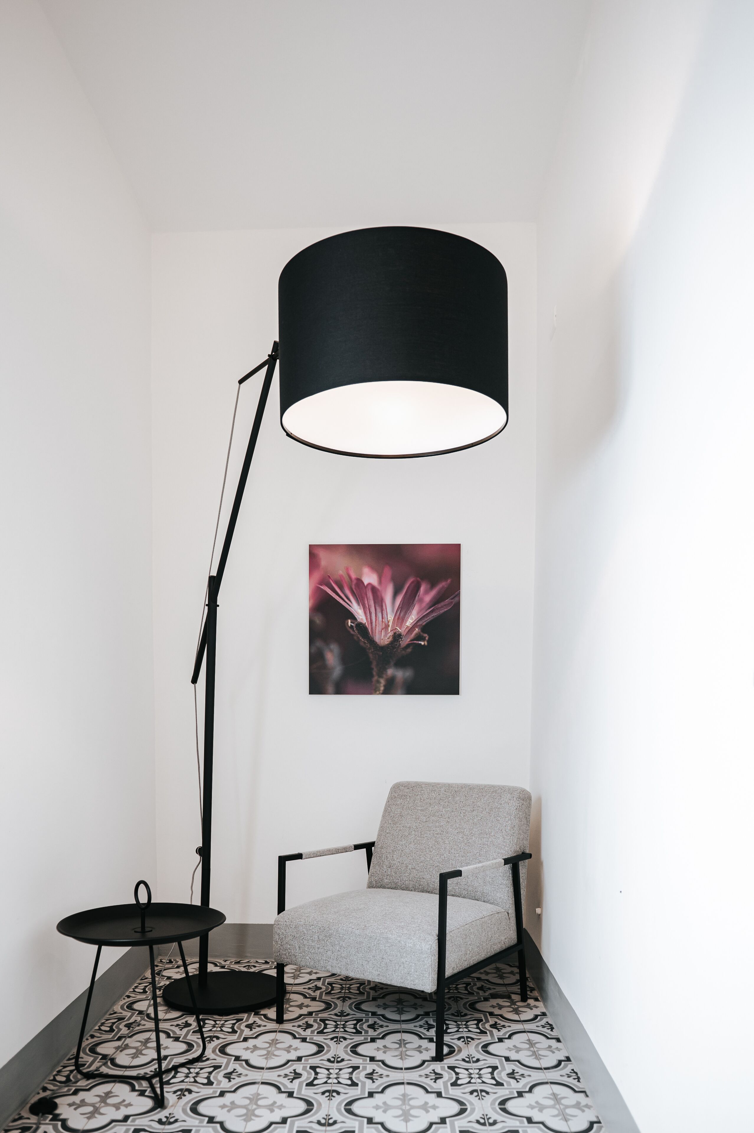 Ein Eckzimmer mit einem gemusterten Boden, einem grauen Sessel, einer schwarzen Stehlampe und einem gerahmten Bild an der Wand.