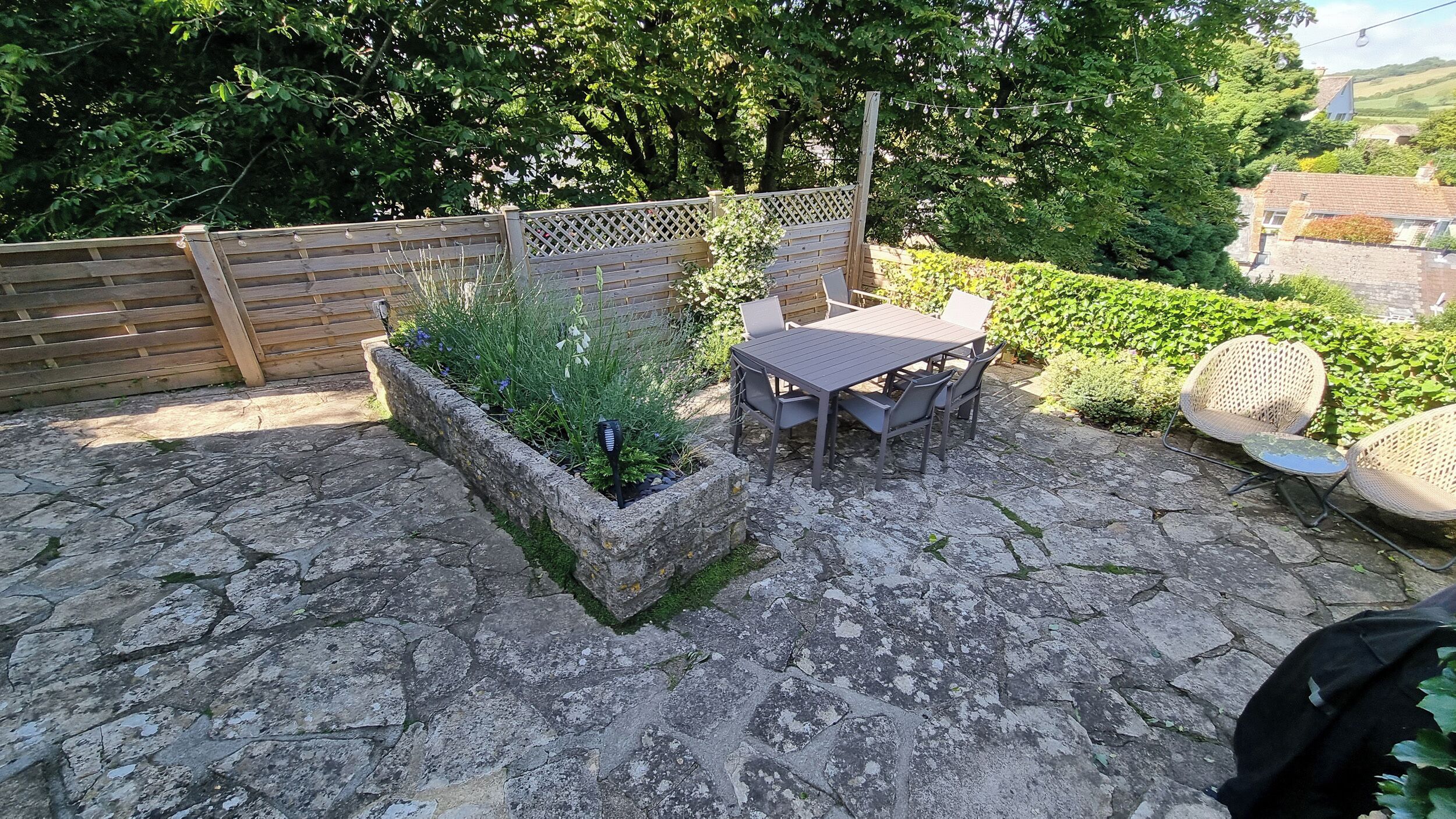 A stone patio with a wooden table and chairs, surrounded by a garden with a wooden fence and greenery.