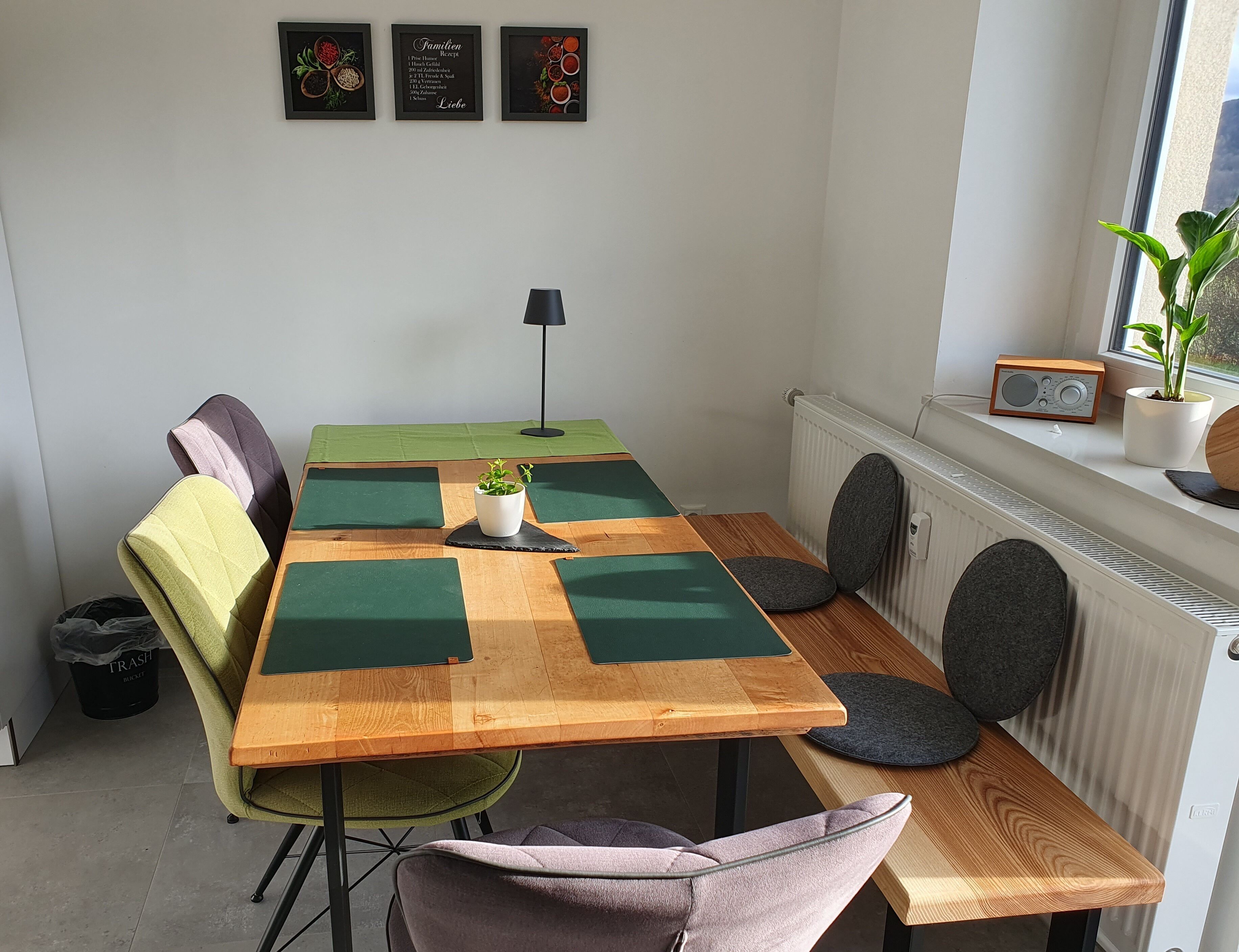 Un coin repas avec une table en bois, des chaises vertes et violettes, une plante en pot et un radiateur.
