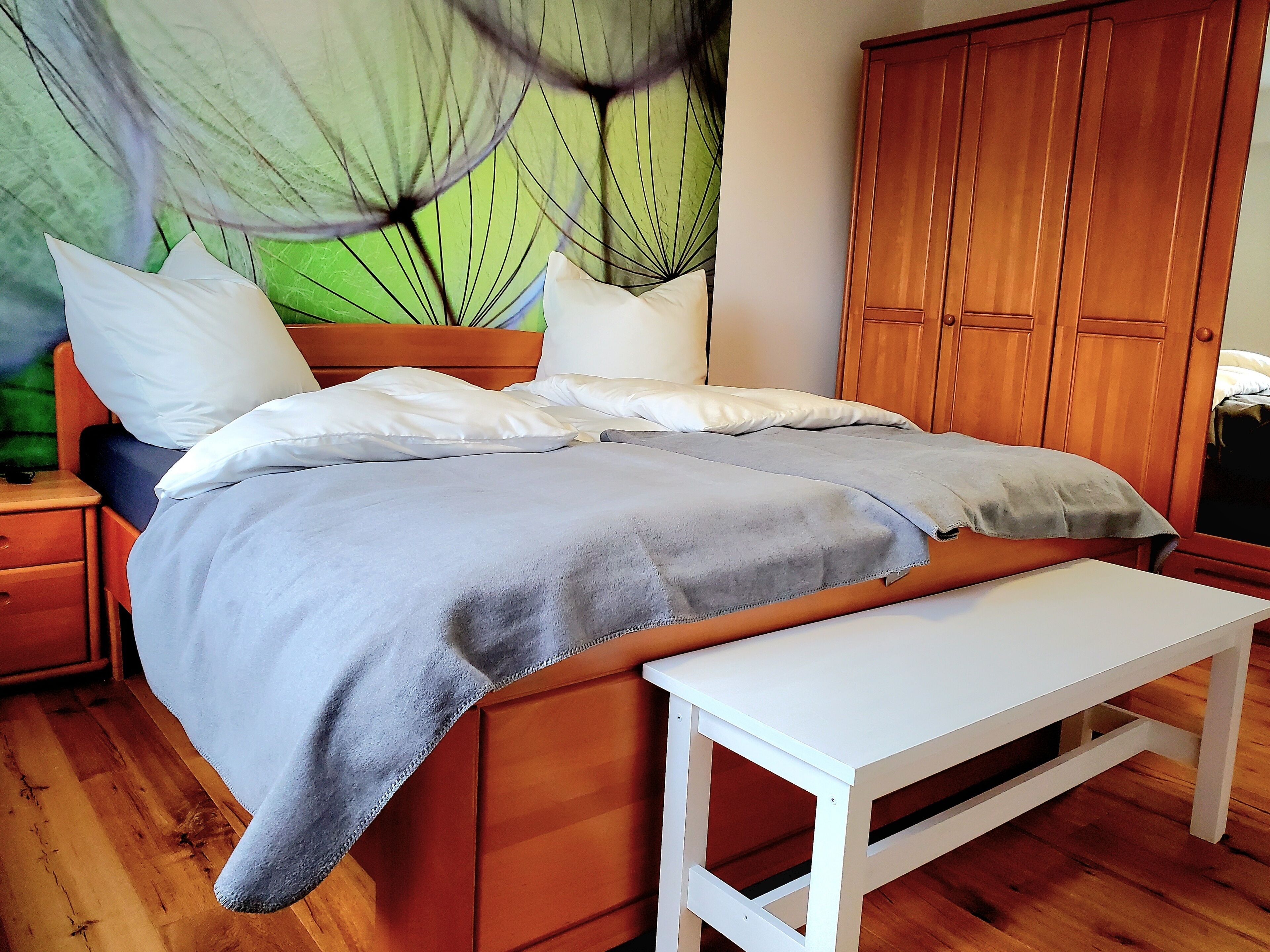 A bed with a grey blanket, white pillows, and a wooden headboard. There is a wooden wardrobe to the right and a white bench in front of the bed.