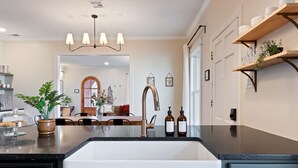 Una cocina con encimera negra, lavavajillas de acero inoxidable y pileta blanca. Hay dos botellas en la encimera, una planta en maceta y un estante de madera con vajilla.