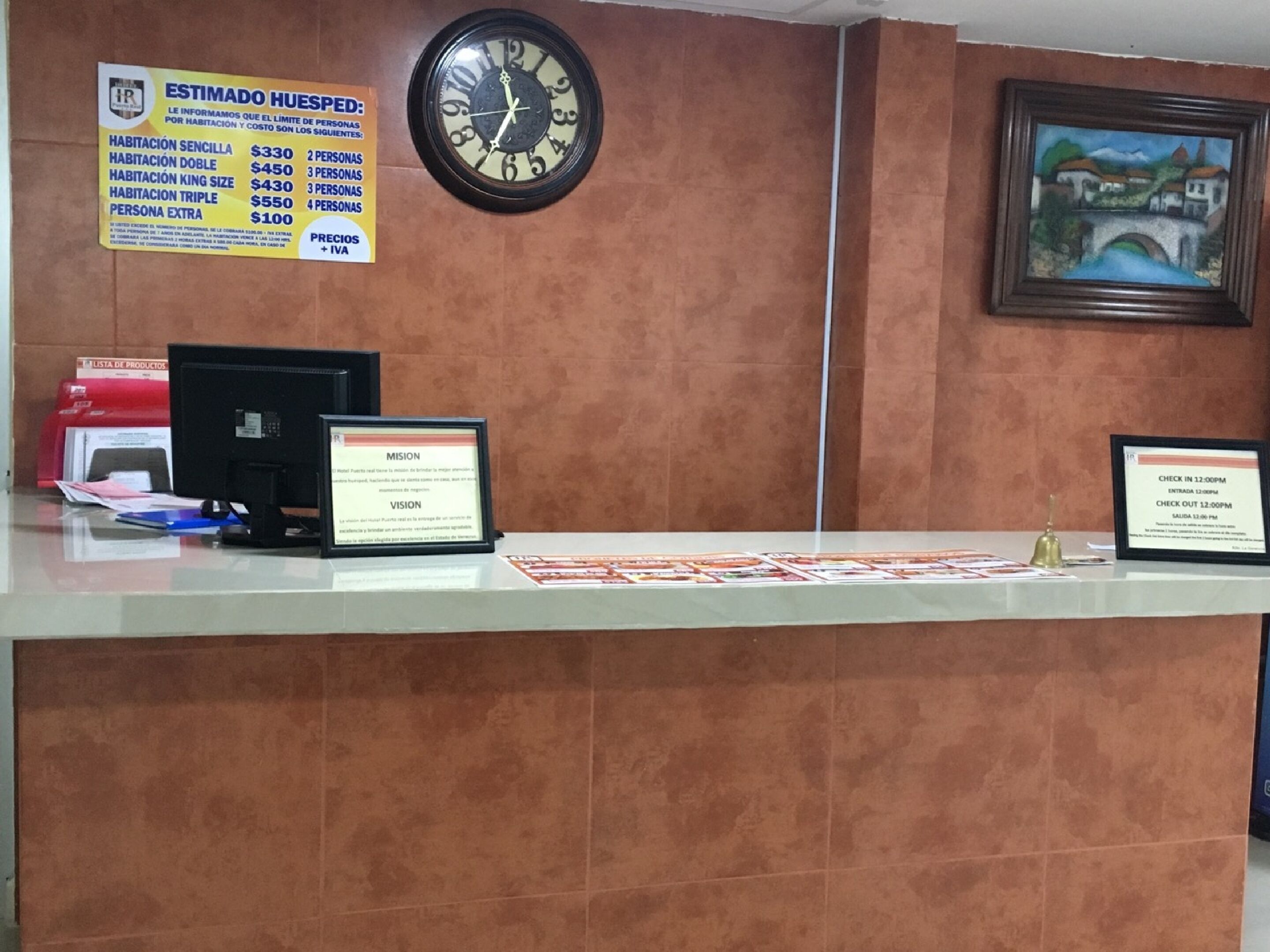 A hotel reception desk with a clock, a framed painting, and informational signs about room rates and check-in/check-out times.