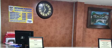 A hotel reception desk with a clock, a framed painting, and informational signs about room rates and check-in/check-out times.