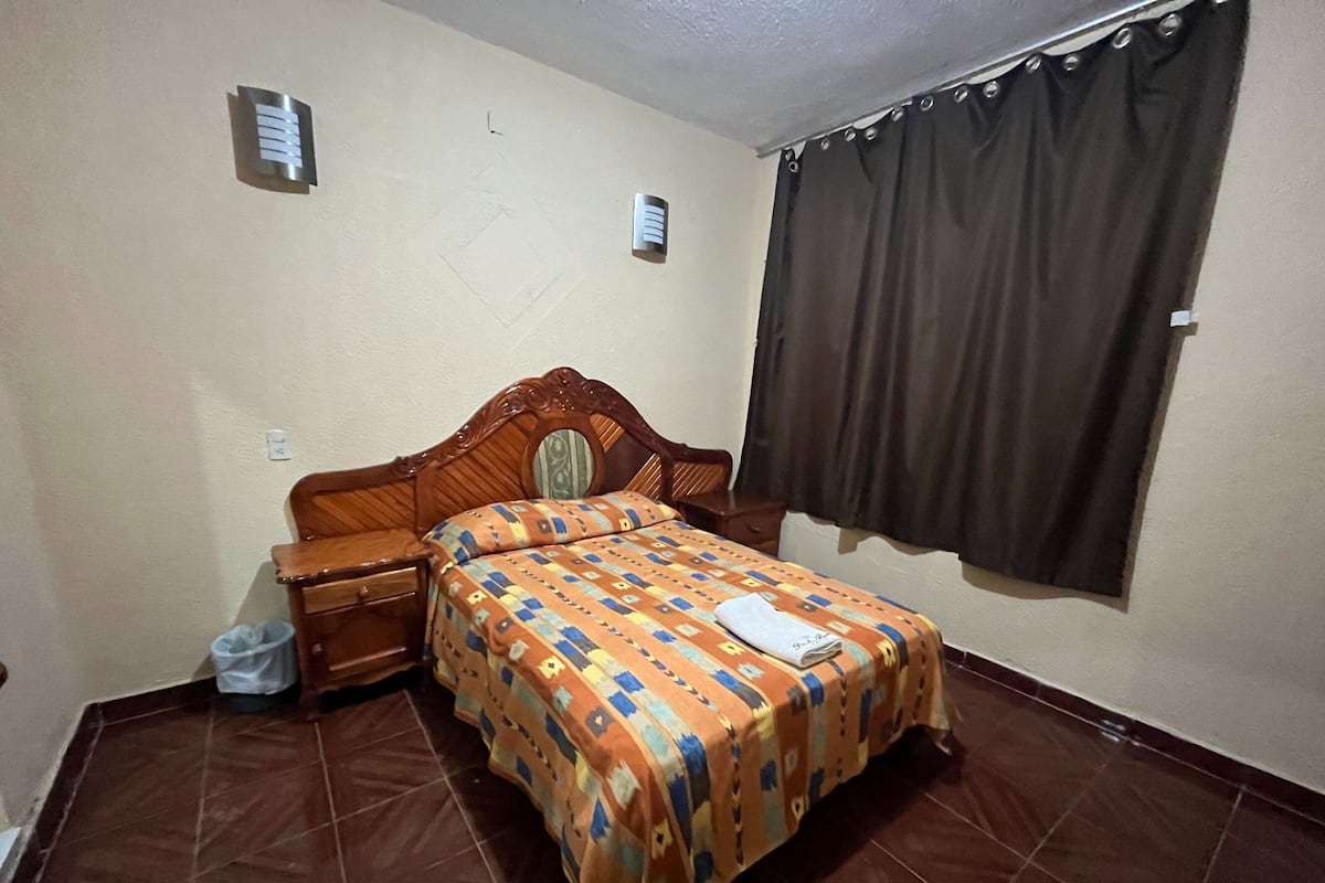 A bedroom with a wooden bed, a nightstand, a bedside table, a patterned bedspread, a window with curtains, and a tiled floor.