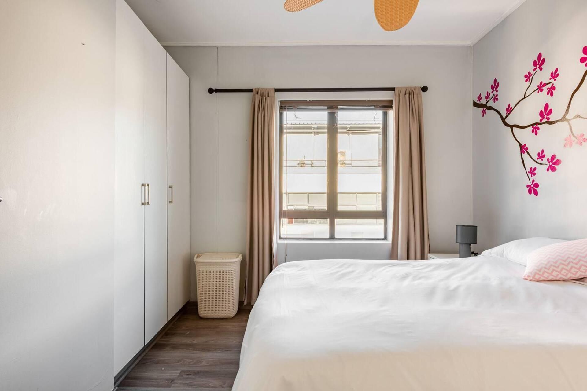 Une chambre à coucher avec un lit, une fenêtre avec des rideaux, une lampe fixée au mur et une poubelle.