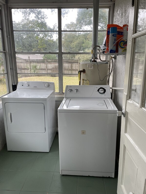 Une buanderie équipée de deux lave-linge et deux sèche-linge à chargement frontal, d’une fenêtre donnant sur un jardin, et d’une étagère où sont rangés les produits de nettoyage.