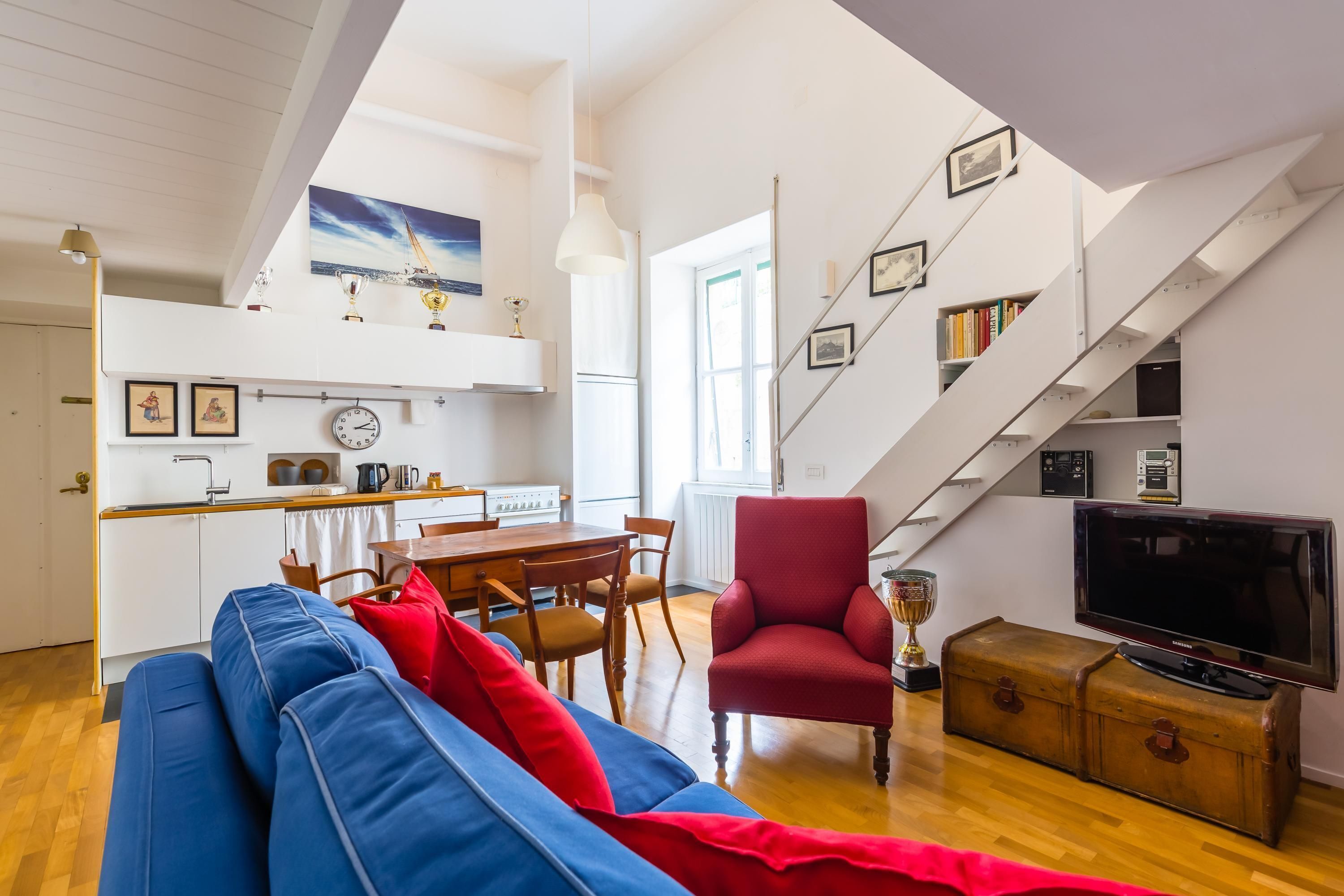 A spacious living area with a blue sofa, red armchair, and a wooden chest of drawers. A staircase with bookshelves is visible in the background.