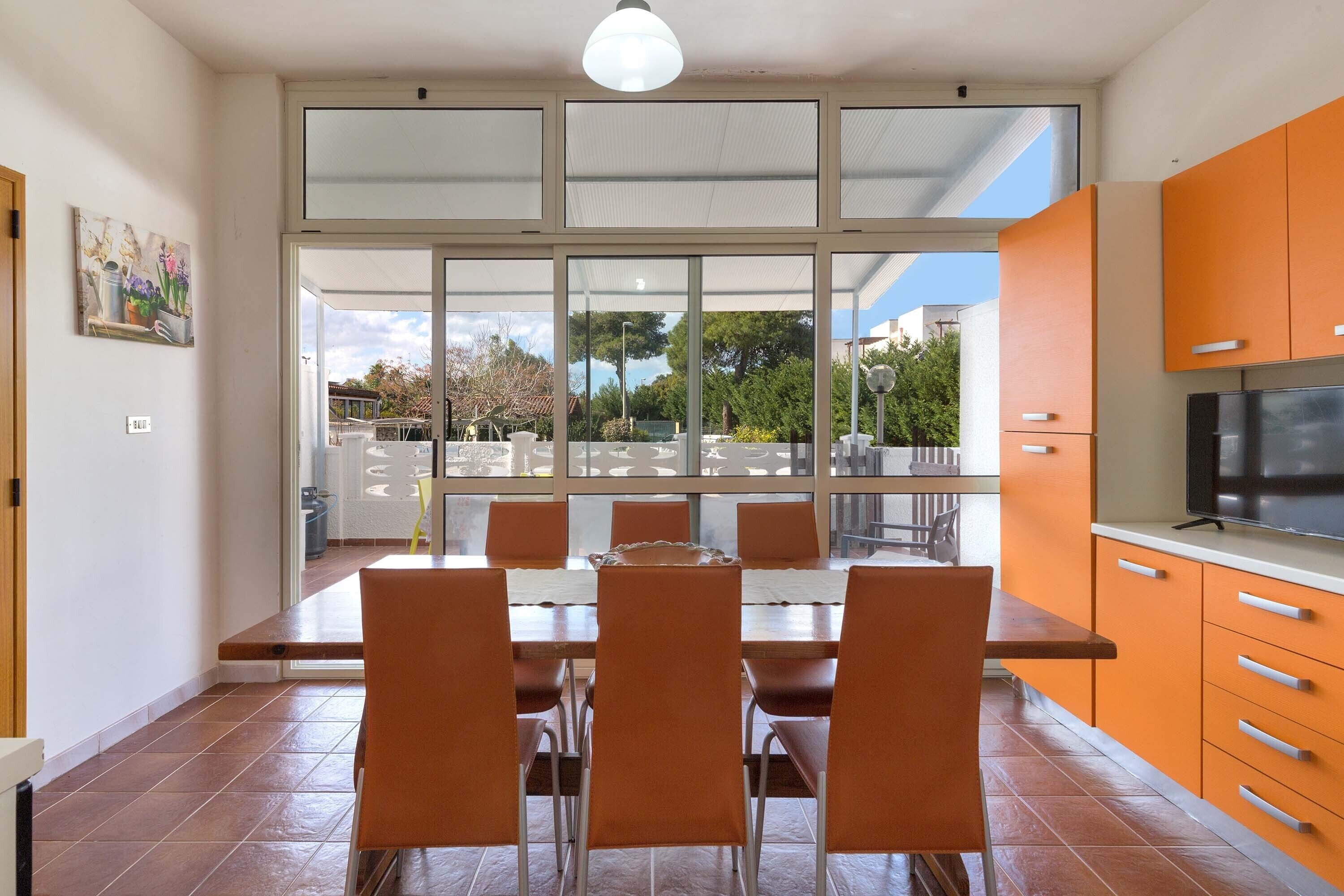 A modern kitchen with orange cabinets, a dining table with chairs, and a large window overlooking a garden.