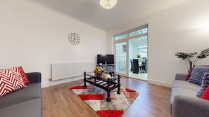 A modern living room with a wooden floor, a coffee table with fruits and wine, a TV, and a large window with a view of a dining area. - Charming 5-bed House in Romford (Romford)