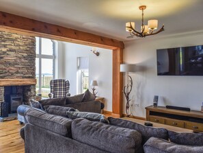 A living room with a stone fireplace, a plaid armchair, a grey sofa, a wooden TV stand, and a chandelier. - Uninterrupted views over Coll Beach and sea views out over Broadbay. (All Outer Hebrides)