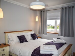 A neatly made bed with white and purple bedding, a wooden headboard, and a window with grey curtains.