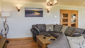 A living room with a sectional sofa, a wooden coffee table, a painting, wall sconces, and a wooden door.