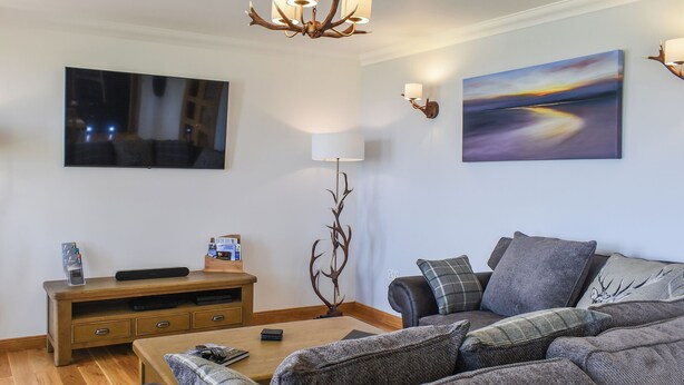A modern living room with a flat-screen TV, a wooden entertainment unit, a sofa with multiple cushions, a coffee table, and a chandelier. - Uninterrupted views over Coll Beach and sea views out over Broadbay. (All Outer Hebrides)