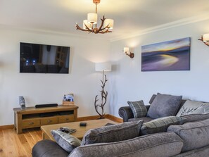 Un salon moderne doté d’un téléviseur à écran plat, d’un meuble de rangement en bois, d’un canapé agrémenté de plusieurs coussins, d’une table basse et d’un lustre.