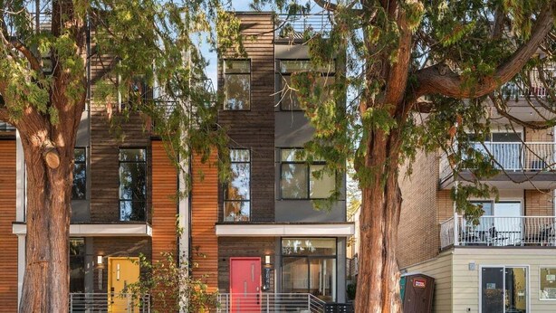 A multi-story residential building with a modern design, featuring a mix of wood and metal materials, large windows, and a visible balcony on the upper floor.