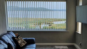 A room with a dark blue sofa, a window with blinds, a TV on the wall, and a view of a lake and mountains.