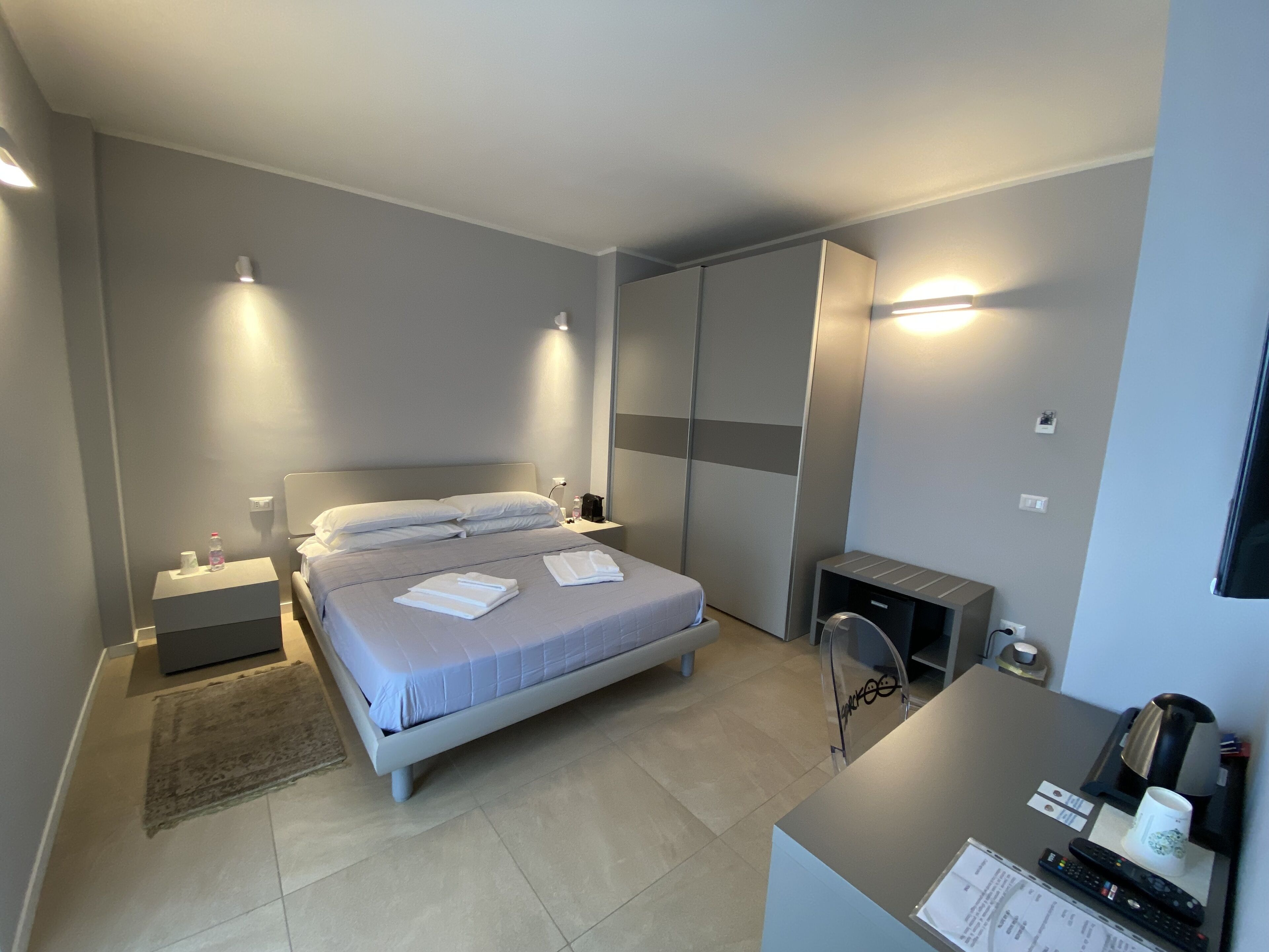 A modern hotel room with a large bed, bedside tables, a desk with a chair, and a wardrobe.