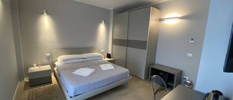 A modern hotel room with a large bed, bedside tables, a desk with a chair, and a wardrobe.