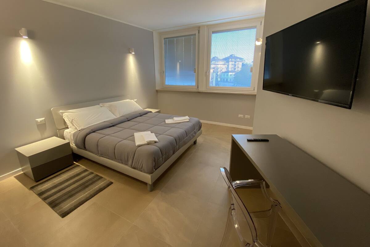 Une chambre moderne avec un lit, des tables de chevet, un téléviseur et une fenêtre donnant sur des immeubles.