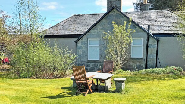 A stone building with a grassy yard, a wooden table, and two chairs.