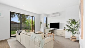 Een moderne woonkamer met een grote schuifpui, een beige bank, een flatscreen televisie en een potplant.