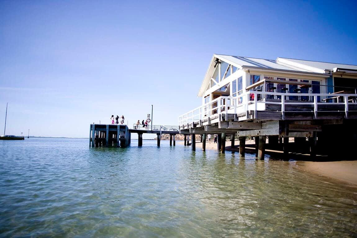 Un edificio frente al agua con un muelle que se extiende sobre el agua, un embarcadero y un pequeño bote amarrado cerca.