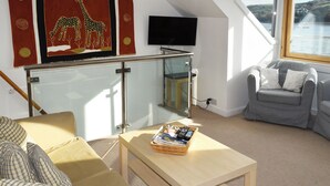 Uma sala de estar com sofá, uma mesa com revista, TV na parede e janela com vista para a água.