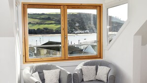 Quarto com duas poltronas cinzas, uma janela ampla com vista para a paisagem costeira e uma parede branca.