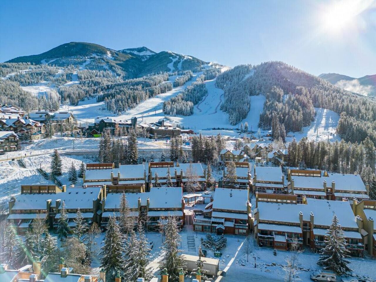 A snow-covered resort with multiple chalet-style buildings, a ski slope in the background, and a mountainous landscape.