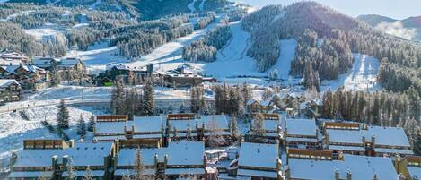A snow-covered resort with multiple chalet-style buildings, a ski slope in the background, and a mountainous landscape.
