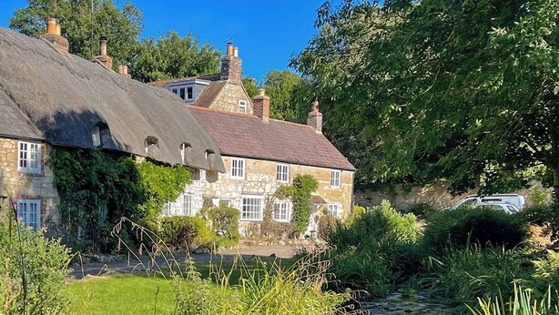 A stone house with a thatched roof, a small pond, and lush greenery.