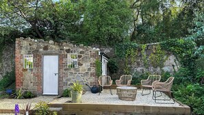 A rustic stone building with a white door, a small window, and a garden area with wicker chairs and a round table.