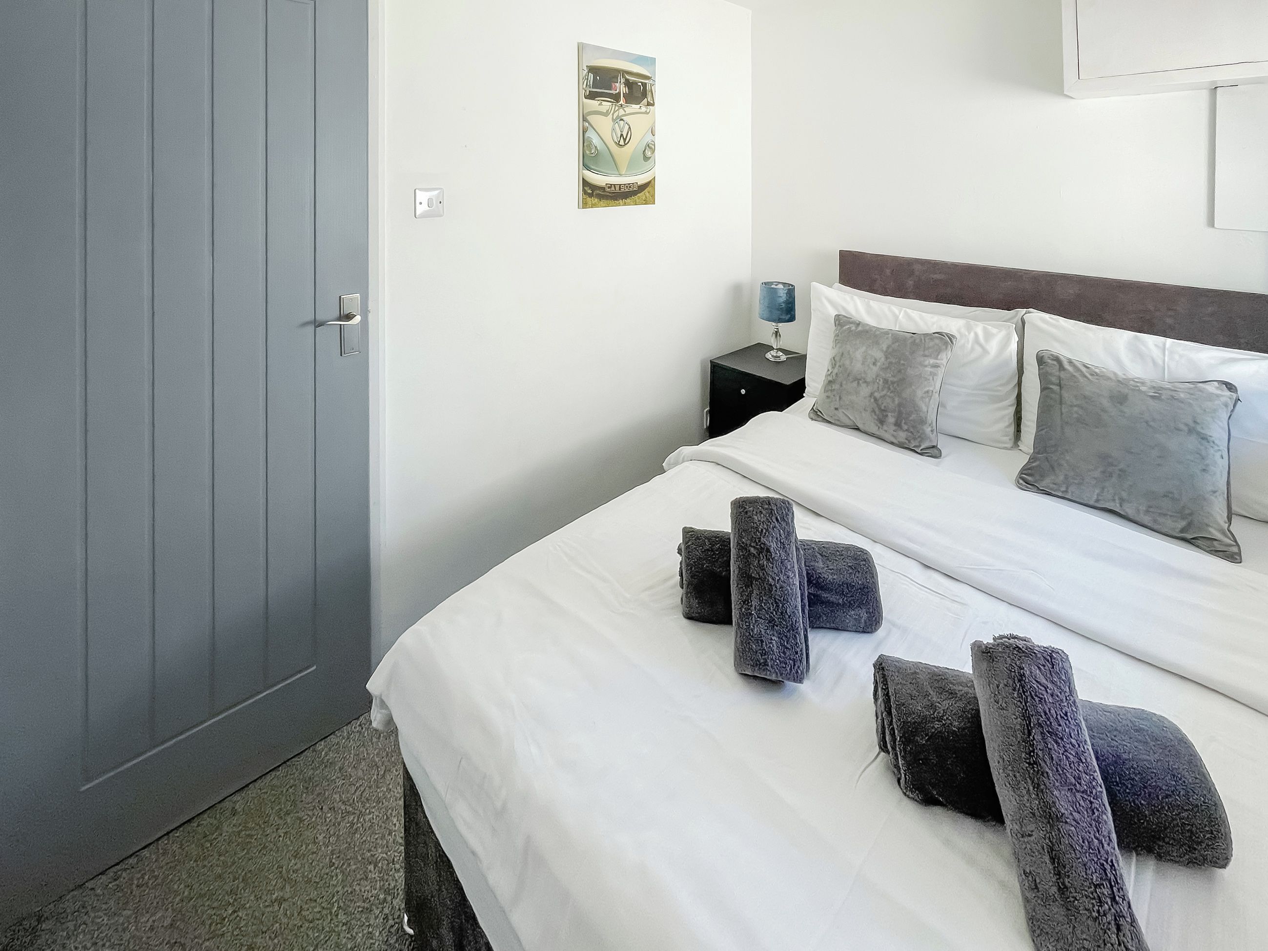 A hotel room with a bed, pillows, a nightstand, a lamp, and a door.