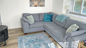 A grey sectional sofa with patterned cushions, a blue patterned rug, a painting of a beach scene, and a window with blinds. - 2 bedroom accommodation in Levens, near Kendal (Levens, near Kendal)