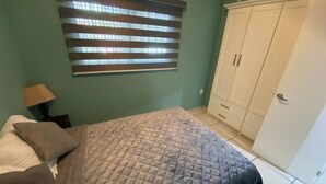 A bedroom with a bed, a nightstand, a lamp, a wardrobe, and a window with blinds. - Condos Tiara (Mazatlán)